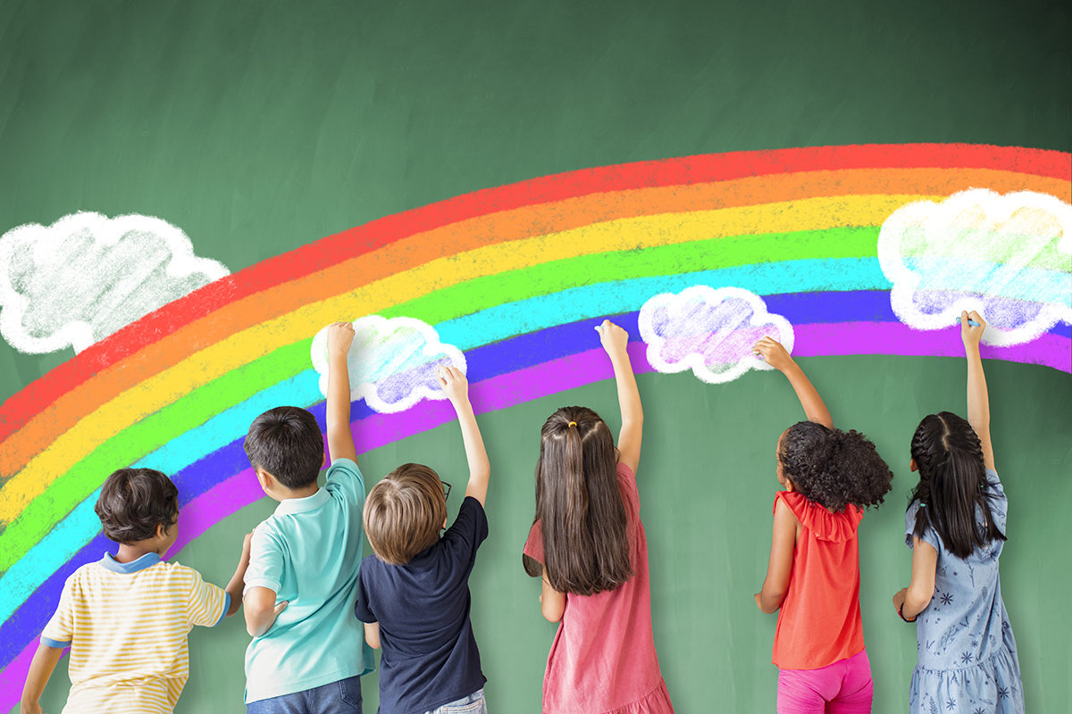 6 children stand with their back to camera drawing with chalk. They have drawn a rainbow with clouds on a teal coloured wall.